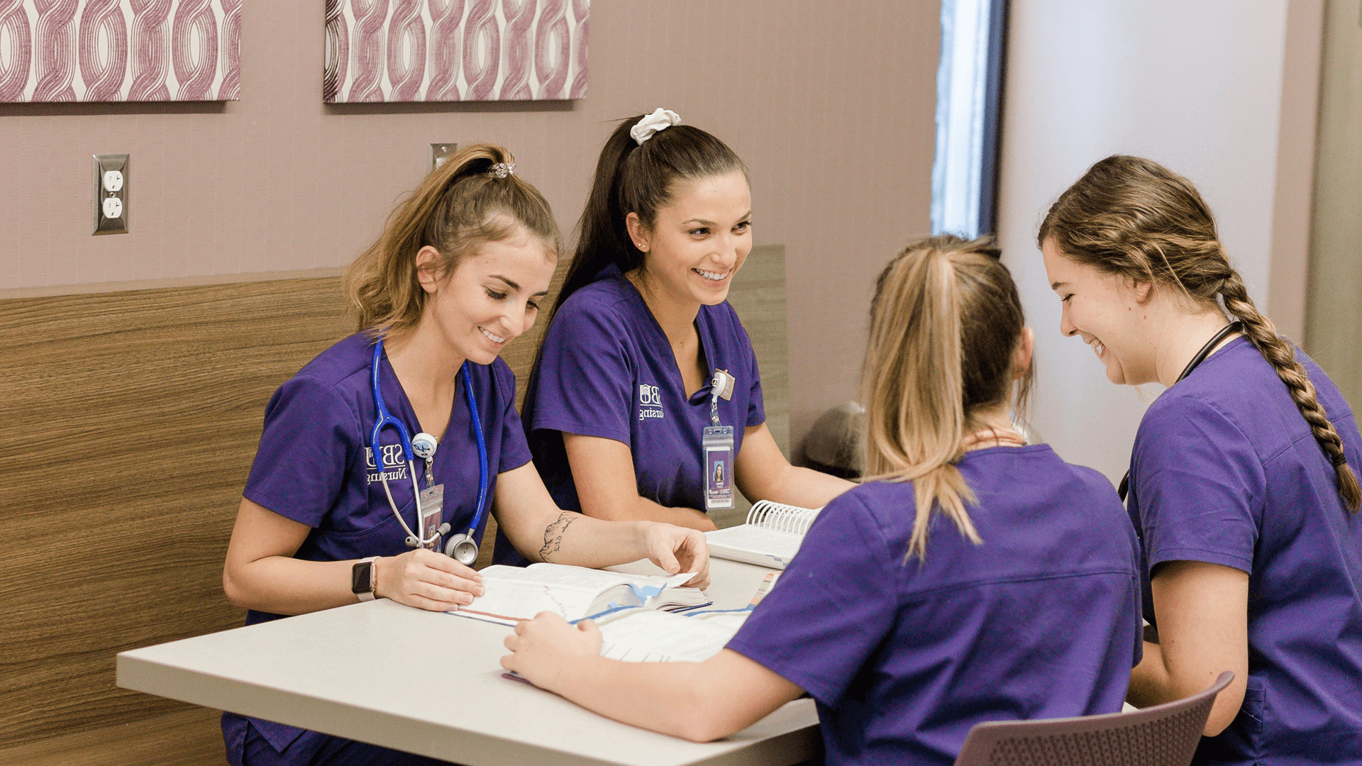 four girls wearing purple scrubs smile and laugh as they sit at a table studying together
